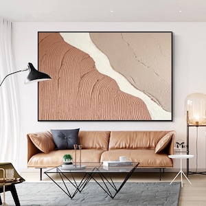 May include: Abstract painting with a textured, layered design in shades of pink and white. The painting is framed in black and hangs on a white wall above a brown leather sofa.