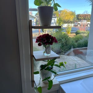 May include: A wooden shelf unit with three shelves, mounted on a window frame. The top shelf holds a potted plant with green leaves, the middle shelf holds a glass vase with red flowers, and the bottom shelf holds a potted plant with green leaves.