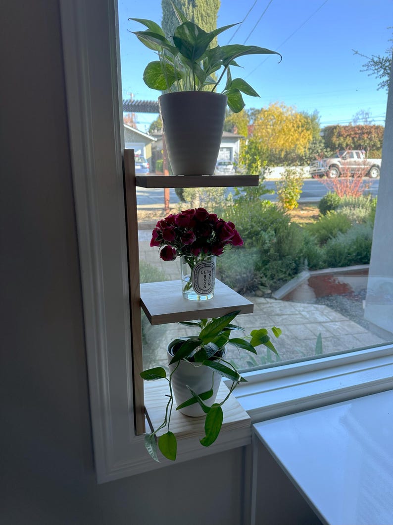 May include: A wooden shelf unit with three shelves, mounted on a window frame. The top shelf holds a potted plant with green leaves, the middle shelf holds a glass vase with red flowers, and the bottom shelf holds a potted plant with green leaves.