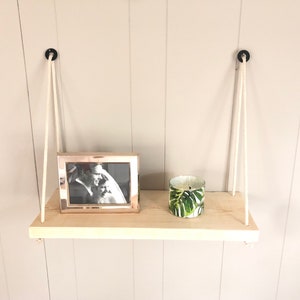 May include: A wooden shelf with white ropes hanging from the ceiling. The shelf is holding a framed black and white photo and a candle with a green tropical leaf design.