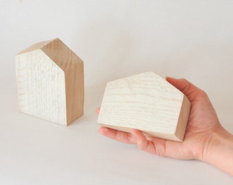 May include: Two wooden house-shaped blocks, one larger than the other, with a whitewashed finish. The smaller block is being held in a hand.
