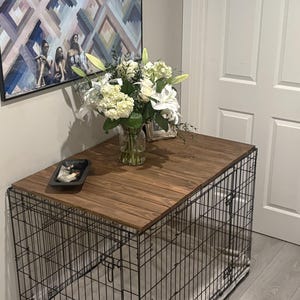 Peut inclure: Une cage pour chien avec un dessus en bois fonc&eacute;, surmont&eacute;e d'un vase de fleurs blanches. La cage est en fil de fer noir et se trouve dans une pi&egrave;ce avec un tableau et une porte blanche.