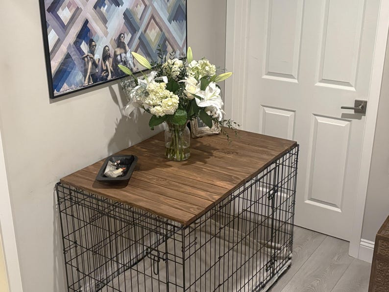 Peut inclure: Une cage pour chien avec un dessus en bois fonc&eacute;, surmont&eacute;e d'un vase de fleurs blanches. La cage est en fil de fer noir et se trouve dans une pi&egrave;ce avec un tableau et une porte blanche.