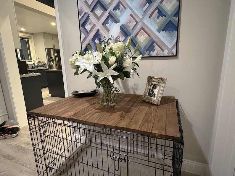 Peut inclure: Un enclos pour chien avec un dessus en bois, orn&eacute; d'un bouquet de fleurs blanches dans un vase en verre, d'une photo encadr&eacute;e et d'un bol noir. Une &oelig;uvre d'art g&eacute;om&eacute;trique est accroch&eacute;e au mur.