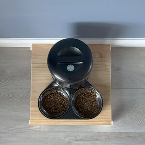 May include: Black automatic pet feeder with two bowls filled with brown pet food. The feeder is on a wooden platform.