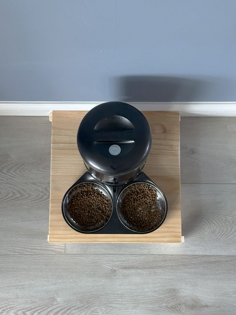 May include: Black automatic pet feeder with two bowls filled with brown pet food. The feeder is on a wooden platform.