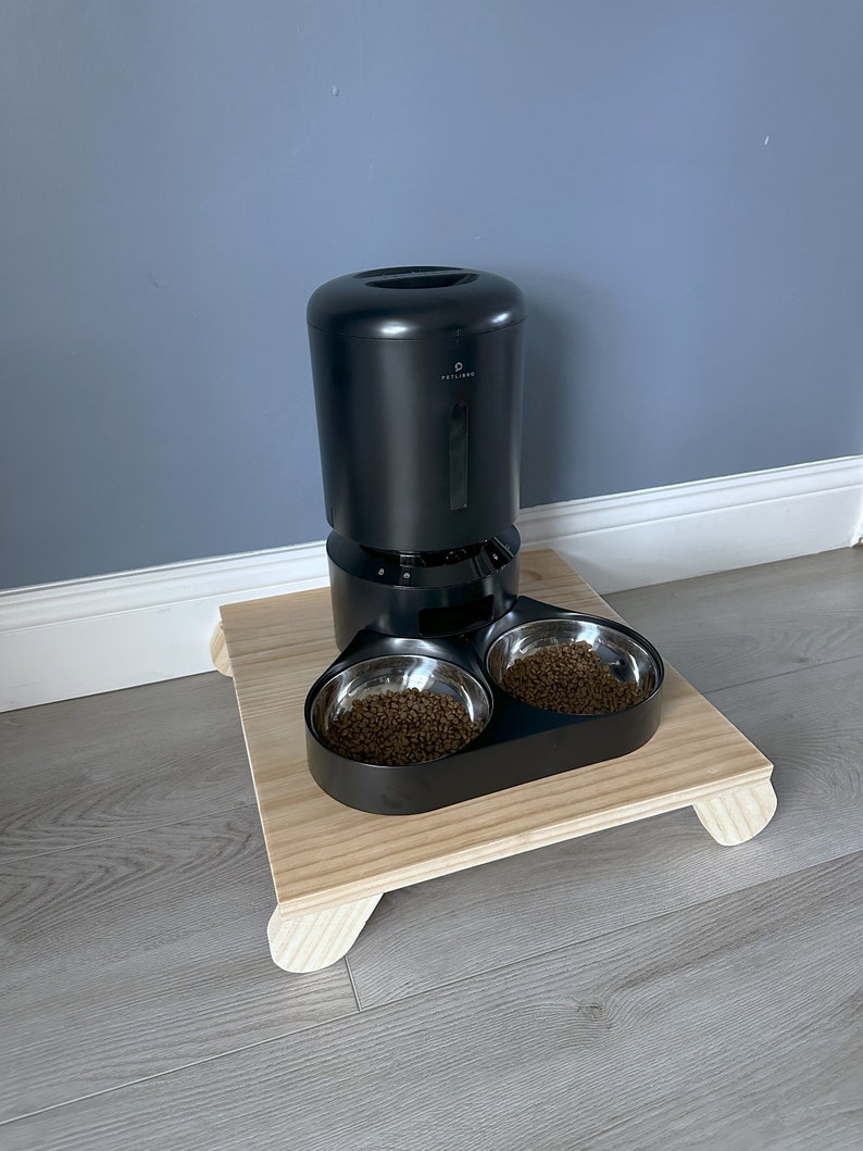 May include: Black automatic pet feeder with two stainless steel bowls filled with brown pet food. The feeder is on a wooden platform with rounded legs.