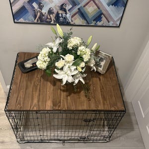 Peut inclure: Une cage pour chien d&eacute;corative avec un dessus en bois fonc&eacute; et une base en cage en m&eacute;tal noir. Le dessus est orn&eacute; d'une composition florale de lys blancs, de roses et d'hortensias. Une photo encadr&eacute;e et un plateau noir sont &eacute;galement sur le dessus.