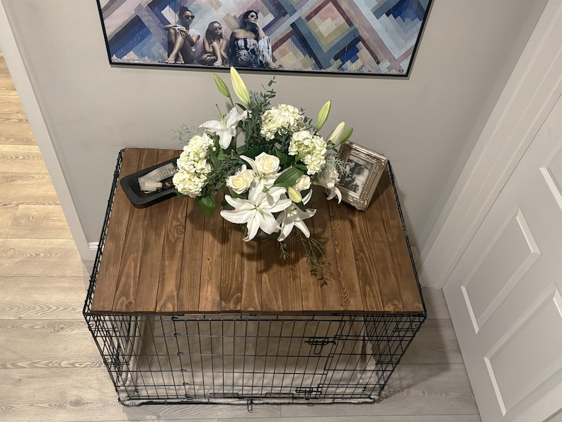 Peut inclure: Une cage pour chien d&eacute;corative avec un dessus en bois fonc&eacute; et une base en cage en m&eacute;tal noir. Le dessus est orn&eacute; d'une composition florale de lys blancs, de roses et d'hortensias. Une photo encadr&eacute;e et un plateau noir sont &eacute;galement sur le dessus.