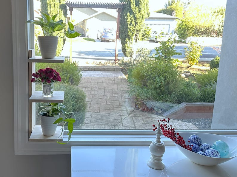 May include: A white wooden shelf unit with three shelves, each holding a potted plant. The top shelf has a green plant, the middle shelf has a bouquet of red flowers, and the bottom shelf has a green plant with trailing vines. The shelf unit is mounted on a white wall next to a window.