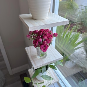 May include: Three wooden shelves mounted on a window, with a potted plant on the top shelf, a vase of pink roses on the middle shelf, and a potted plant on the bottom shelf.