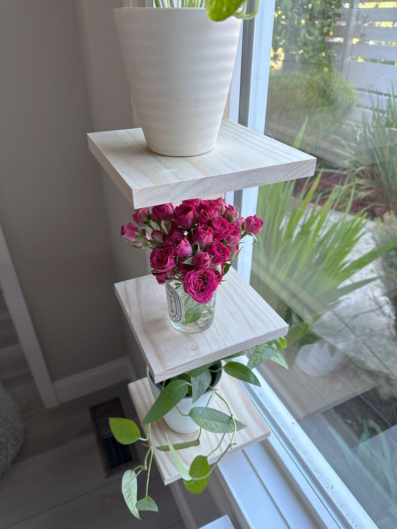 May include: Three wooden shelves mounted on a window, with a potted plant on the top shelf, a vase of pink roses on the middle shelf, and a potted plant on the bottom shelf.