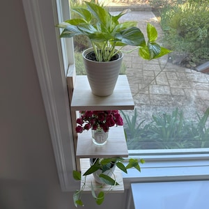 May include: A three-tiered wooden shelf unit mounted on a windowsill, holding three potted plants. The top shelf has a large green plant with variegated leaves in a white ceramic pot. The middle shelf has a small bouquet of pink flowers in a clear glass jar. The bottom shelf has a small green plant with trailing vines in a white ceramic pot.