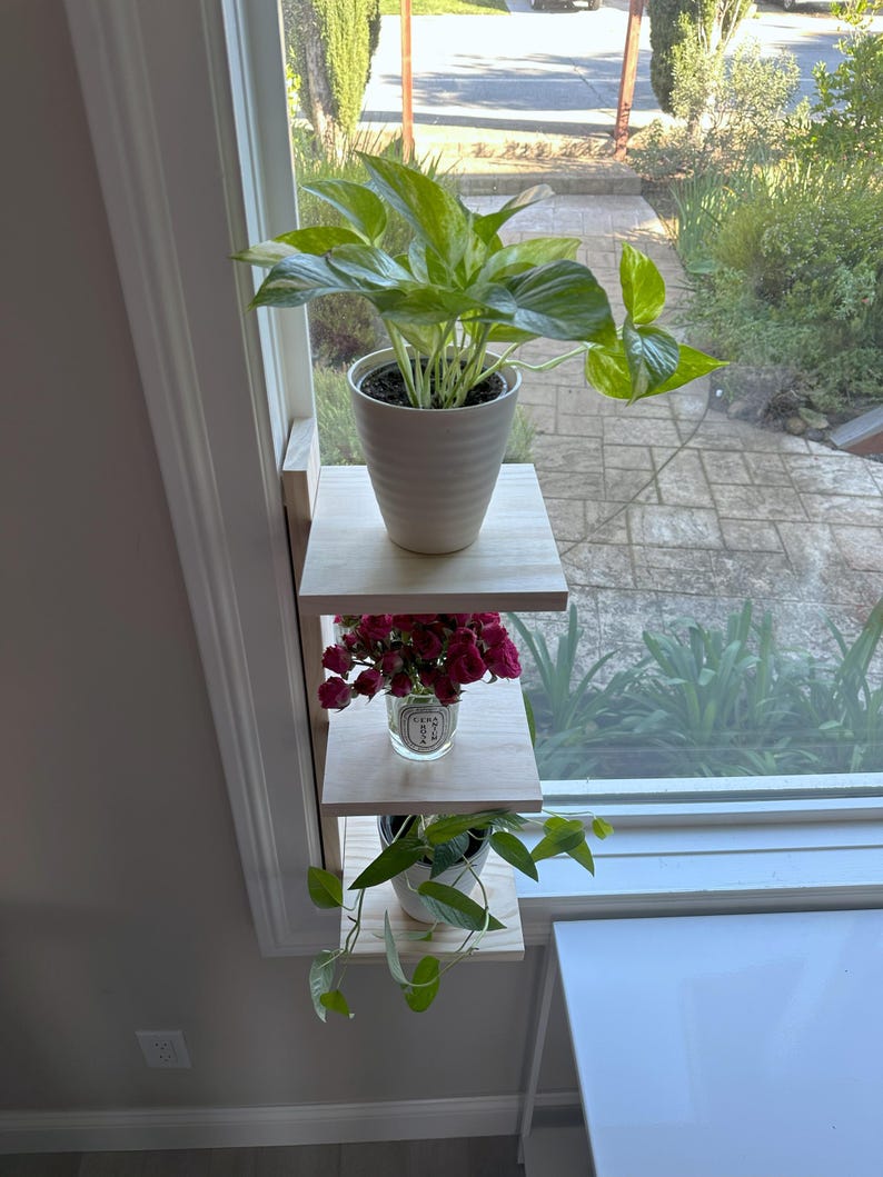 May include: A three-tiered wooden shelf unit mounted on a windowsill, holding three potted plants. The top shelf has a large green plant with variegated leaves in a white ceramic pot. The middle shelf has a small bouquet of pink flowers in a clear glass jar. The bottom shelf has a small green plant with trailing vines in a white ceramic pot.
