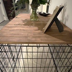 Peut inclure: Une cage pour chien rustique avec un dessus en bois. Le dessus est en bois teint&eacute; et de forme rectangulaire. La base de la cage est en fil de fer noir. Un cadre photo d&eacute;coratif et un vase avec des fleurs sont sur le dessus.