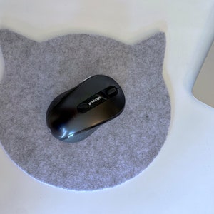 Könnte beinhalten: Ein graues Filz-Mauspad in Form eines Katzenkopfes mit einer schwarzen kabellosen Maus darauf. Die Maus trägt den Schriftzug "Microsoft".