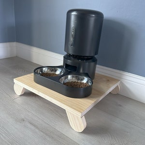 May include: A black automatic pet feeder with two stainless steel bowls on a wooden platform with rounded legs.