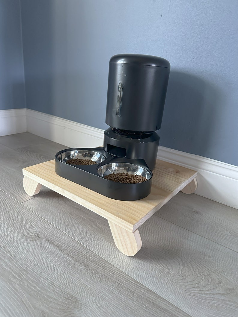 May include: A black automatic pet feeder with two stainless steel bowls on a wooden platform with rounded legs.