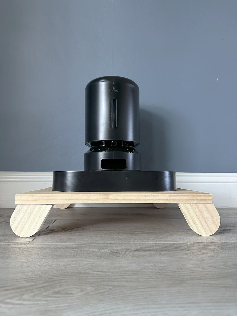 May include: A black pet feeder with a wooden base. The feeder has a round, cylindrical design and a small opening on the front. The base has four rounded legs.
