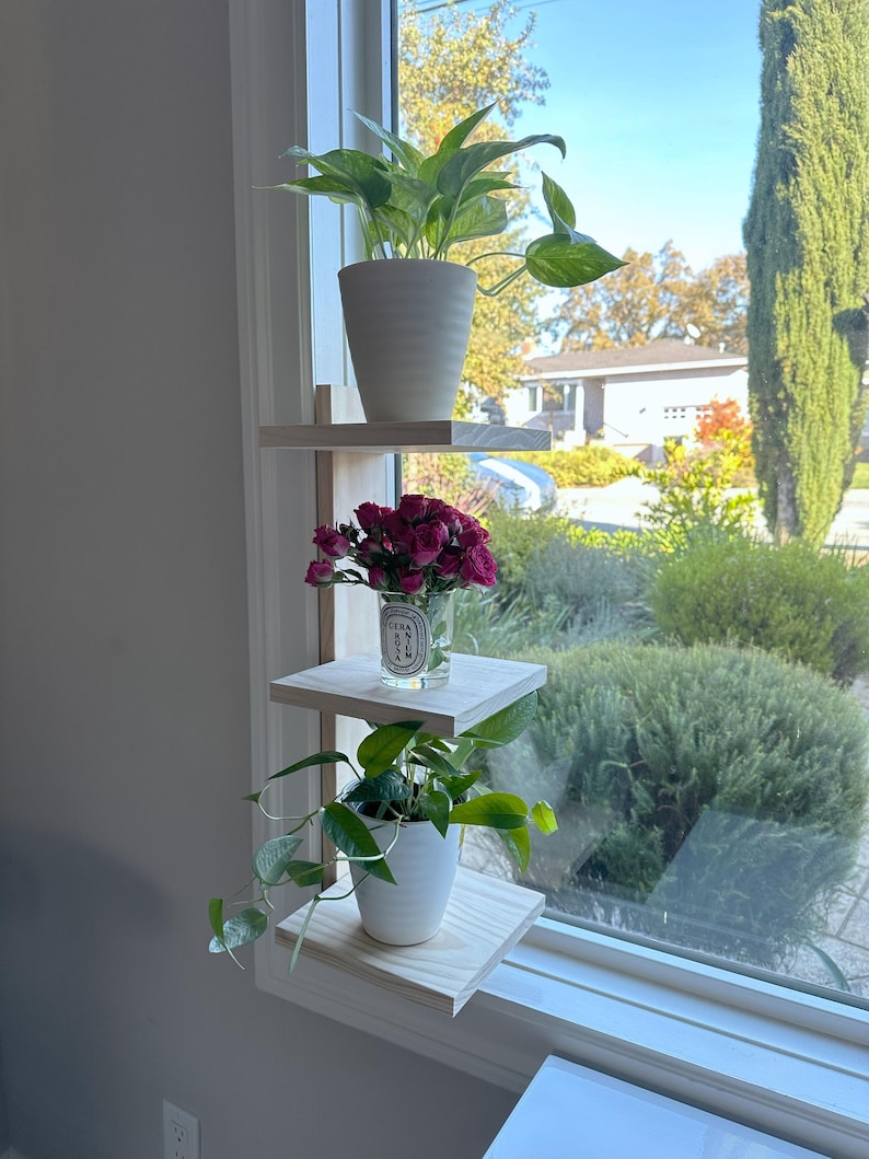 May include: A white wooden shelf unit with three tiers, mounted on a windowsill. The top shelf holds a potted green plant, the middle shelf holds a small glass vase with pink flowers, and the bottom shelf holds a potted green plant.