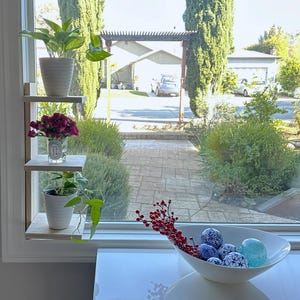 May include: A white wooden shelf unit with three shelves, mounted on a wall next to a window. The shelves are decorated with potted plants and flowers. The window looks out onto a backyard with a patio and green trees.