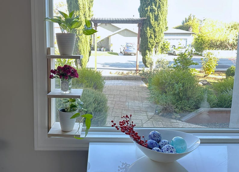 May include: A white wooden shelf unit with three shelves, mounted on a wall next to a window. The shelves are decorated with potted plants and flowers. The window looks out onto a backyard with a patio and green trees.