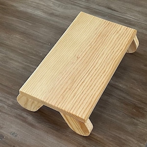 May include: A small, wooden step stool with a natural finish. The stool has a rectangular top and rounded edges. It is made of solid wood and has a simple, rustic design.