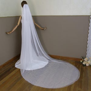 Puede incluir: Un velo de novia de tul blanco con una cola larga. El velo está drapeado sobre un maniquí y fluye por el suelo.