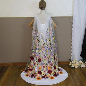 May include: A white wedding dress with a floral embroidered cape. The cape features a variety of colourful flowers, including yellow sunflowers, orange, and red blooms. The dress has a low back and a long train.