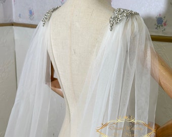 May include: White tulle bridal cape with crystal embellishments on the shoulders. The cape is draped over a dress form.