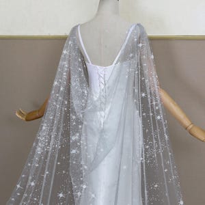 May include: A sheer, floor-length cape made of light gray tulle fabric featuring a white star and sparkle pattern. The cape is draped over a white sleeveless dress with a lace-up back closure, showcasing a celestial design across the fabric.