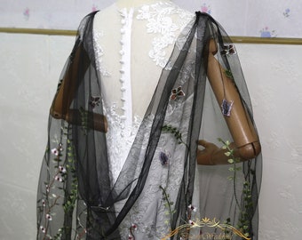Pode incluir: Uma capa de noiva de tule preto com bordados florais e detalhes de borboleta. A capa está drapeada sobre um vestido de noiva de renda branca.