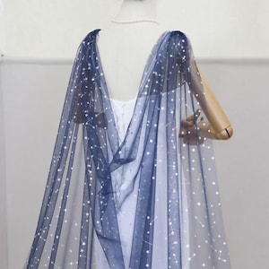 May include: A sheer navy blue tulle cape featuring a pattern of silver stars and constellations. The cape is draped over a white dress on a mannequin, with the fabric extending from the shoulders down to the floor in a flowing, translucent design.