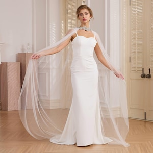 May include: A white tulle bridal cape with a long train. The cape is worn over a white strapless dress.