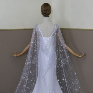 May include: A white tulle wedding cape with a scattered star pattern. The cape is sheer and has a long train.