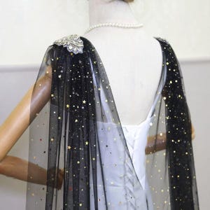 May include: Black tulle cape with gold star accents and jeweled shoulder embellishments. The cape is draped over a white dress, creating a celestial and elegant look. Perfect for special occasions.