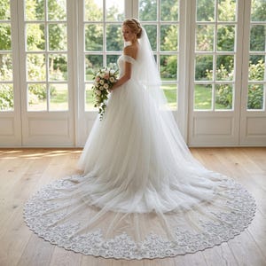 Puede incluir: Vestido de novia blanco con hombros descubiertos y cola larga. El vestido tiene una falda de tul y un velo. La cola tiene un ribete de encaje. La novia está de pie frente a grandes puertas blancas.