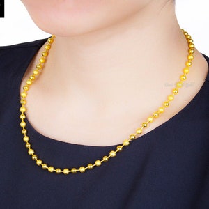 Pu&ograve; includere: Una collana a catena con palline dorate composta da una serie di piccole perle d'oro rotonde e testurizzate collegate tra loro in un filo continuo. La collana &egrave; mostrata su uno sfondo di tessuto scuro, con il testo Best Thai Gold visibile nell'angolo.
