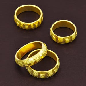 Puede incluir: Cuatro anillos dorados con un patrón geométrico texturizado. Los anillos se muestran sobre una superficie marrón oscuro. Los anillos son de color oro brillante.