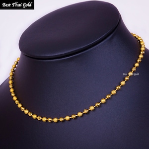 Pu&ograve; includere: Una collana a catena con palline dorate composta da una serie di piccole perle sferiche testurizzate collegate tra loro. La collana &egrave; esposta su un supporto per collo scuro. Il testo "Best Thai Gold" &egrave; visibile in alto a sinistra e a destra.