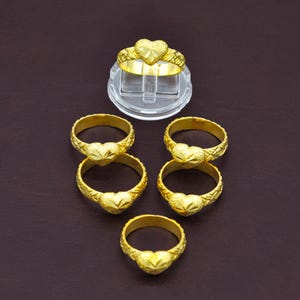 May include: Five gold-toned rings, each featuring a central heart-shaped motif with a faceted, star-like texture. The bands of the rings display a subtle, repeating cross-hatched pattern. One ring is elevated on a clear, circular plastic display stand.