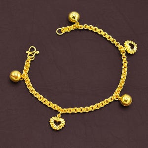 May include: A gold-colored bracelet with a chain design. The bracelet features three small, round charms and two heart-shaped charms. The bracelet is displayed on a dark brown surface.