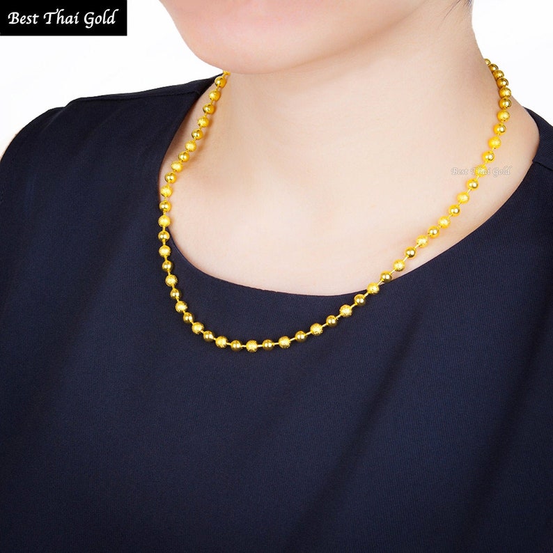 Pu&ograve; includere: Una collana a catena con palline dorate composta da una serie di piccole perle d'oro rotonde e testurizzate collegate tra loro in un filo continuo. La collana &egrave; mostrata su uno sfondo di tessuto scuro, con il testo Best Thai Gold visibile nell'angolo.