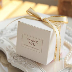 May include: A square white gift box featuring a cream-colored ribbon with gold metallic edges tied in a bow on top. The front of the box displays a rectangular label with the text THANK YOU FOR SHARING THIS MOMENT WITH US in brown lettering.