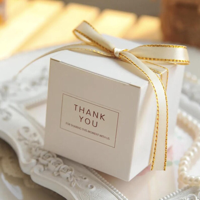 May include: A square white gift box featuring a cream-colored ribbon with gold metallic edges tied in a bow on top. The front of the box displays a rectangular label with the text THANK YOU FOR SHARING THIS MOMENT WITH US in brown lettering.
