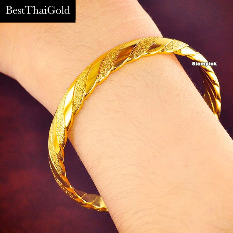 May include: A gold bangle bracelet with a twisted design. The bracelet has a textured surface and is worn on a person's wrist.