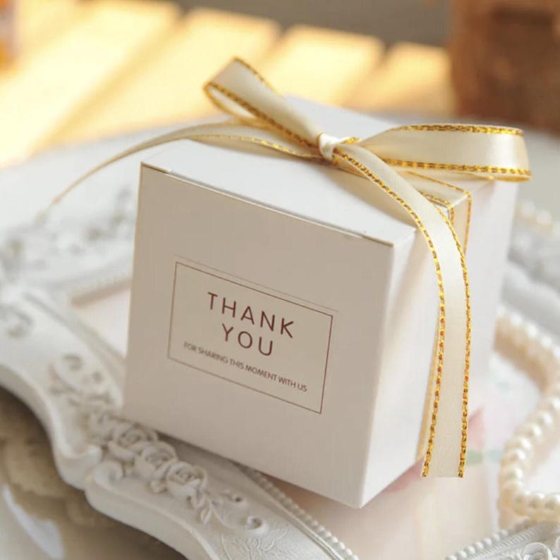 Pu&ograve; includere: Una scatola regalo quadrata bianca con un nastro color crema dai bordi metallici dorati legato a fiocco sulla parte superiore. La parte anteriore della scatola presenta un'etichetta rettangolare con il testo THANK YOU FOR SHARING THIS MOMENT WITH US in marrone.