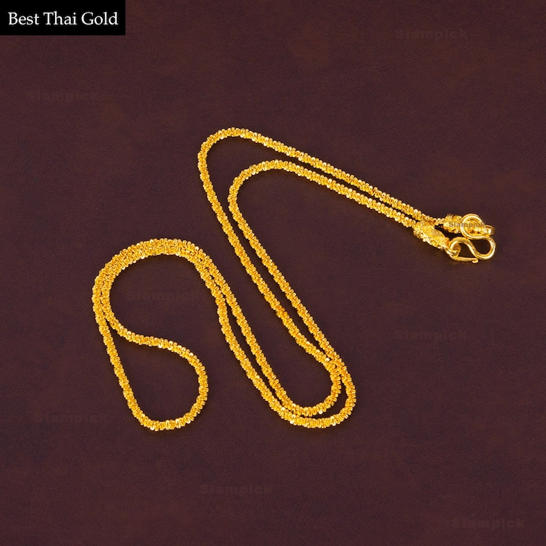 May include: Two gold chains, one longer than the other, are laid out on a dark brown surface. The chains are made of small, linked, round pieces of gold. The chains are both closed with a clasp.