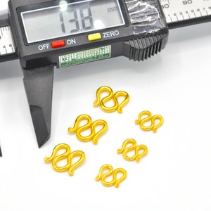 May include: Six gold-colored metal clasps, each shaped like a double loop, are arranged on a white surface. A digital caliper is visible in the background, measuring 1.38 mm.