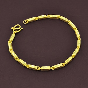 Op de afbeelding: Een goudkleurige armband met een reeks rechthoekige schakels. De schakels zijn verbonden door kleine, afgeronde connectoren. De armband heeft een haaksluiting. De achtergrond is effen donkerbruin.